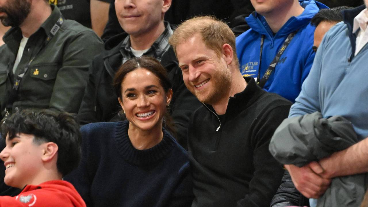 Harry und Meghan posieren auf der Tribüne bei einem Sportevent für ein Foto.