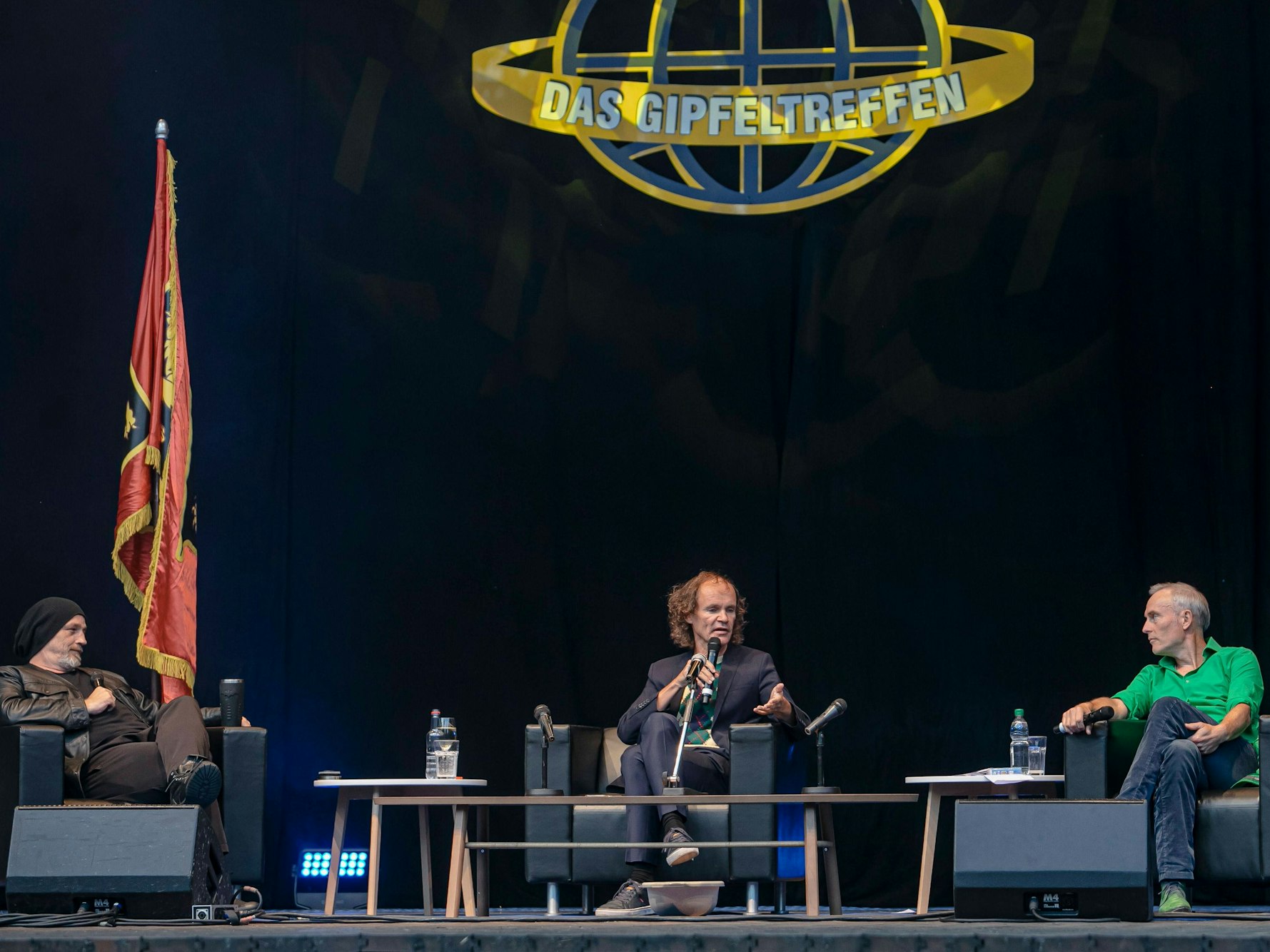 Drei Männer sitzen in niedrigen Sesseln auf einem Podium und diskutieren. Im Hintergrund Flaggen und das Logo "Das Gipfeltreffen".