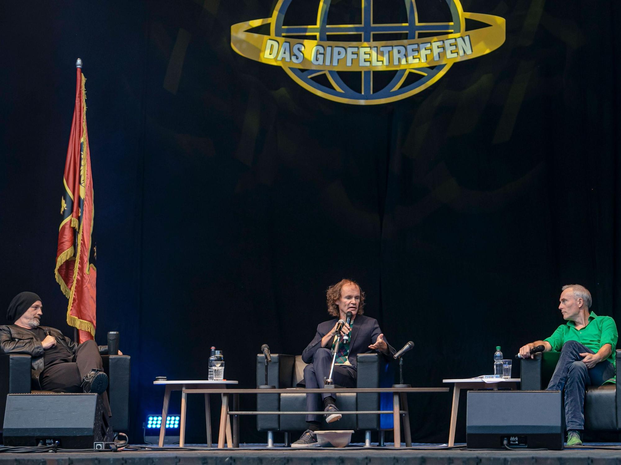 Drei Männer sitzen in niedrigen Sesseln auf einem Podium und diskutieren. Im Hintergrund Flaggen und das Logo "Das Gipfeltreffen".