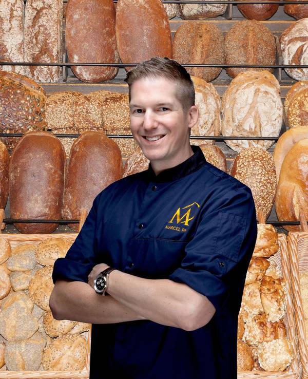 Frische Brote und Brötchen im Regal einer Bäckerei, davo steht ein junger Mann in blauer Bäckerjacke und lächelt