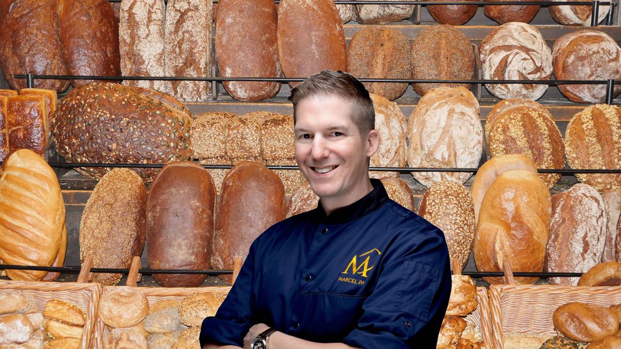Frische Brote und Brötchen im Regal einer Bäckerei, davo steht ein junger Mann in blauer Bäckerjacke und lächelt