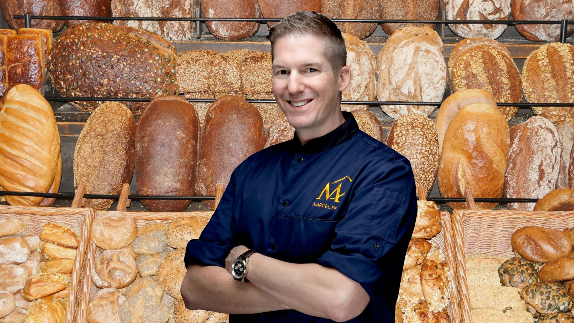 Frische Brote und Brötchen im Regal einer Bäckerei, davo steht ein junger Mann in blauer Bäckerjacke und lächelt