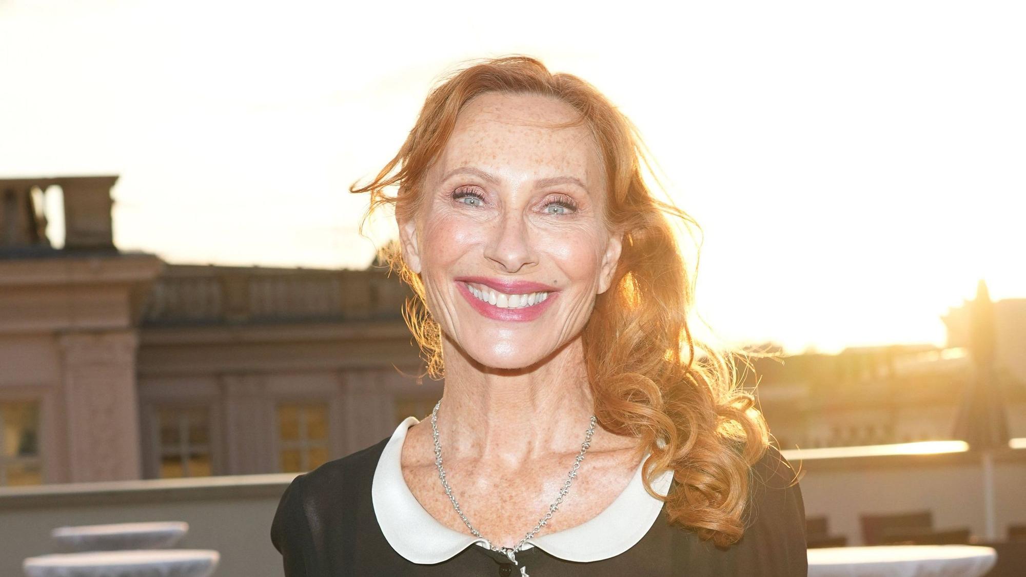 Eine rothaarige Frau lacht in die Kamera, im Hintergrund strahlt die Sonne über Hausdächern.