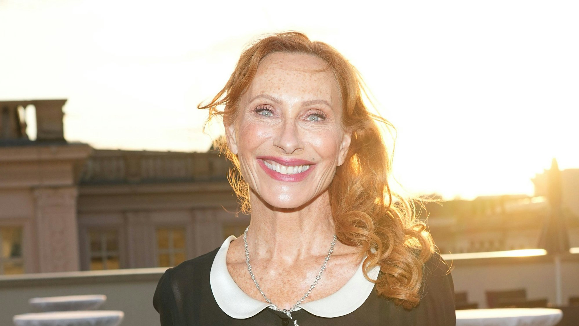 Eine rothaarige Frau lacht in die Kamera, im Hintergrund strahlt die Sonne über Hausdächern.