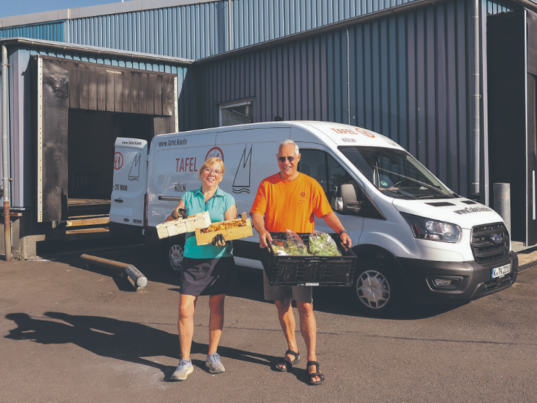 Ein Mann und eine Frau stehen vor einem weißen Ford Transit und halten Kisten mit Lebensmitteln in den Händen.