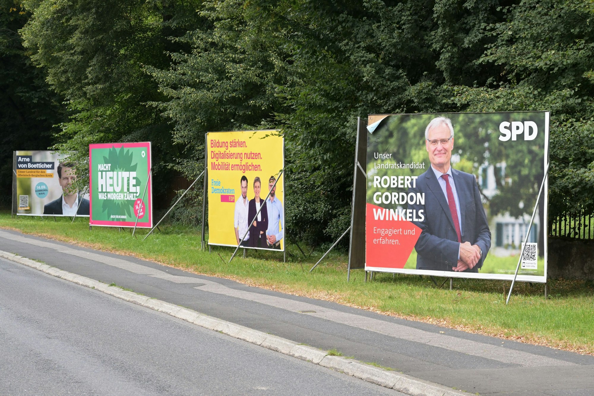 Plakate der CDU, Grünen, FDP und SPD stehen nebeneinander auf der Overather Straße.