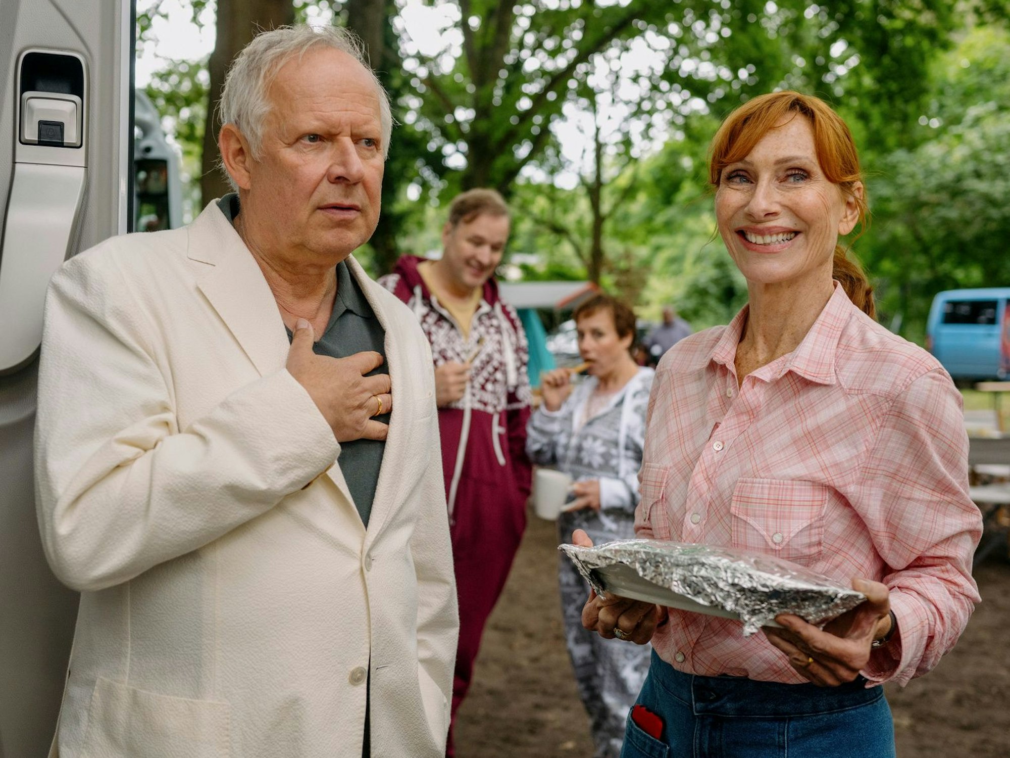 Ein Mann im hellen Anzug und eine Frau in schicker Bluse stehen mit einem Kuchenpaket auf einem Campingplatz.