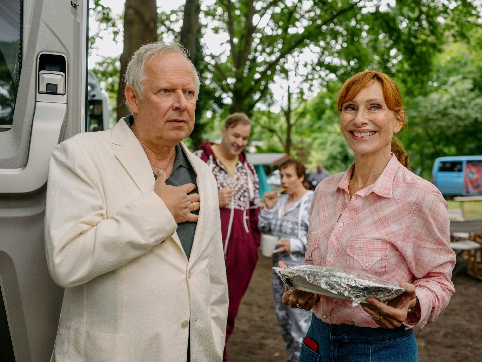 Ein Mann und eine Frau stehen vor einem Wohnmobil unter einem Baum. Der Mann trägt einen hellen Anzug, die Frau hält ein mit Alufolie umwickeltes Paket in der Hand.