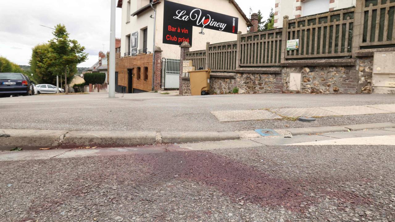 Auf einer Straße ist ein großer Blutfleck, im Hintergrund das Gebäude mit einer Werbung für eine Bar.