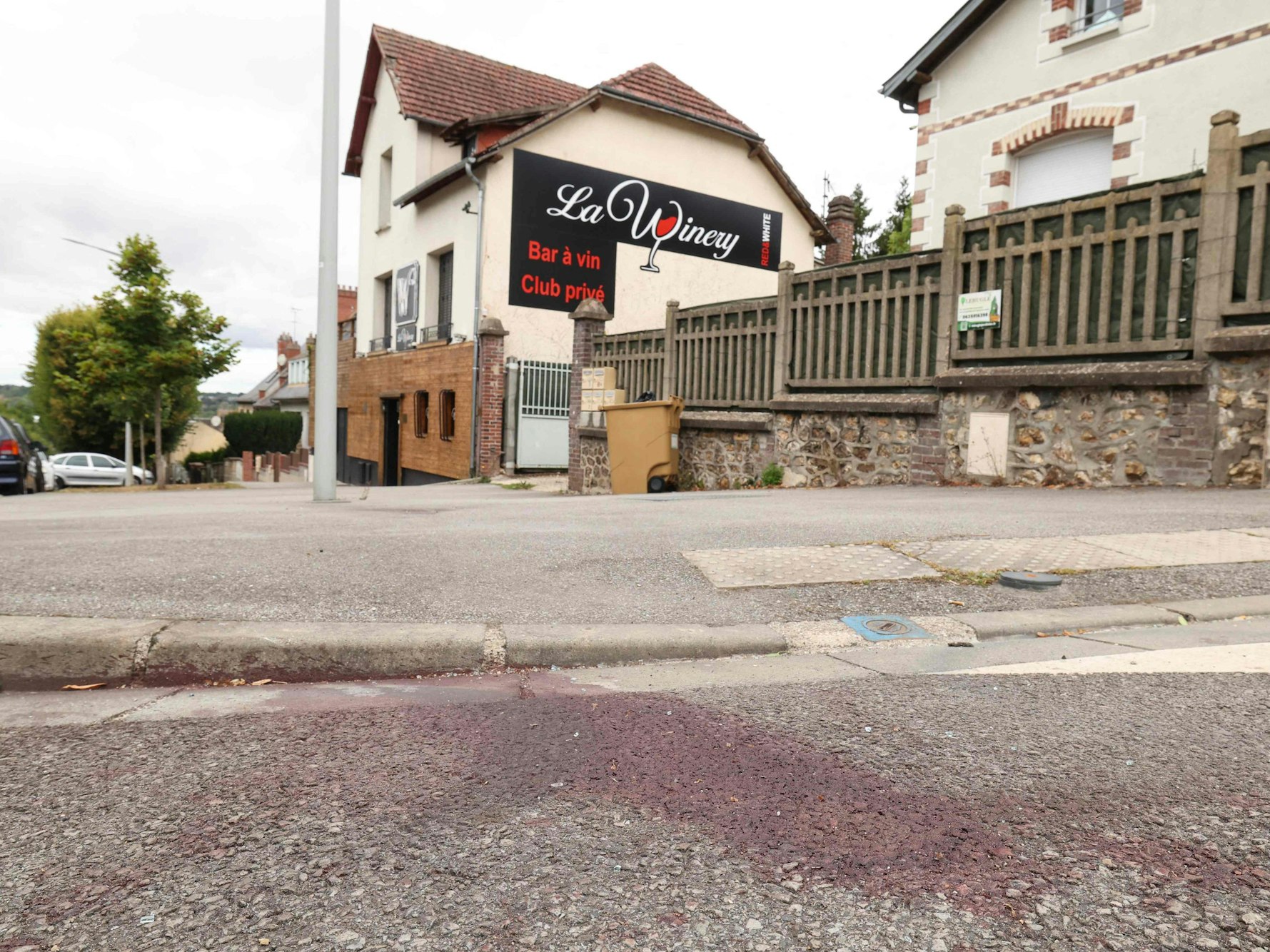 Auf einer Straße ist ein großer Blutfleck, im Hintergrund das Gebäude mit einer Werbung für eine Bar.