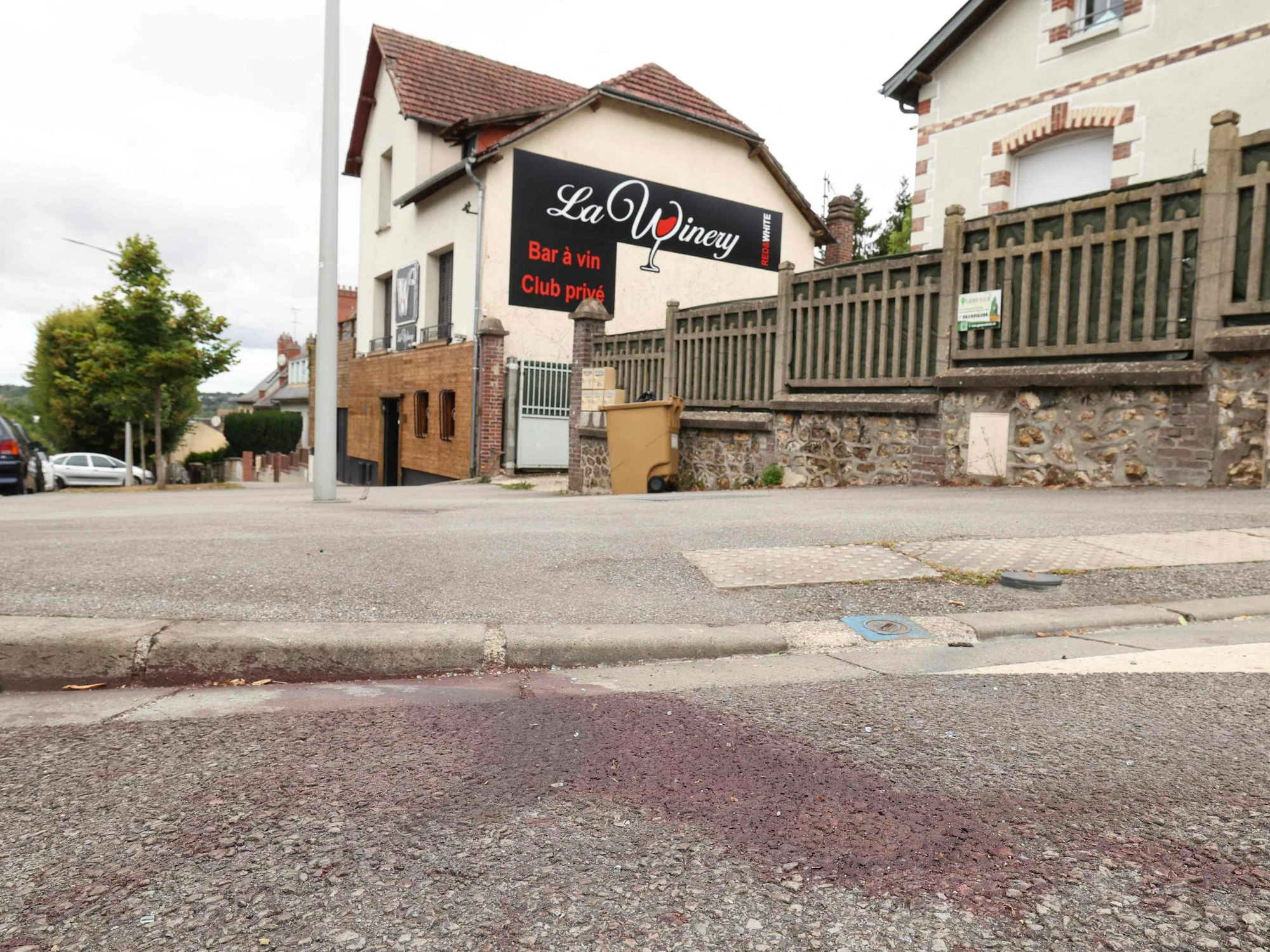 Auf einer Straße ist ein großer Blutfleck, im Hintergrund das Gebäude mit einer Werbung für eine Bar.