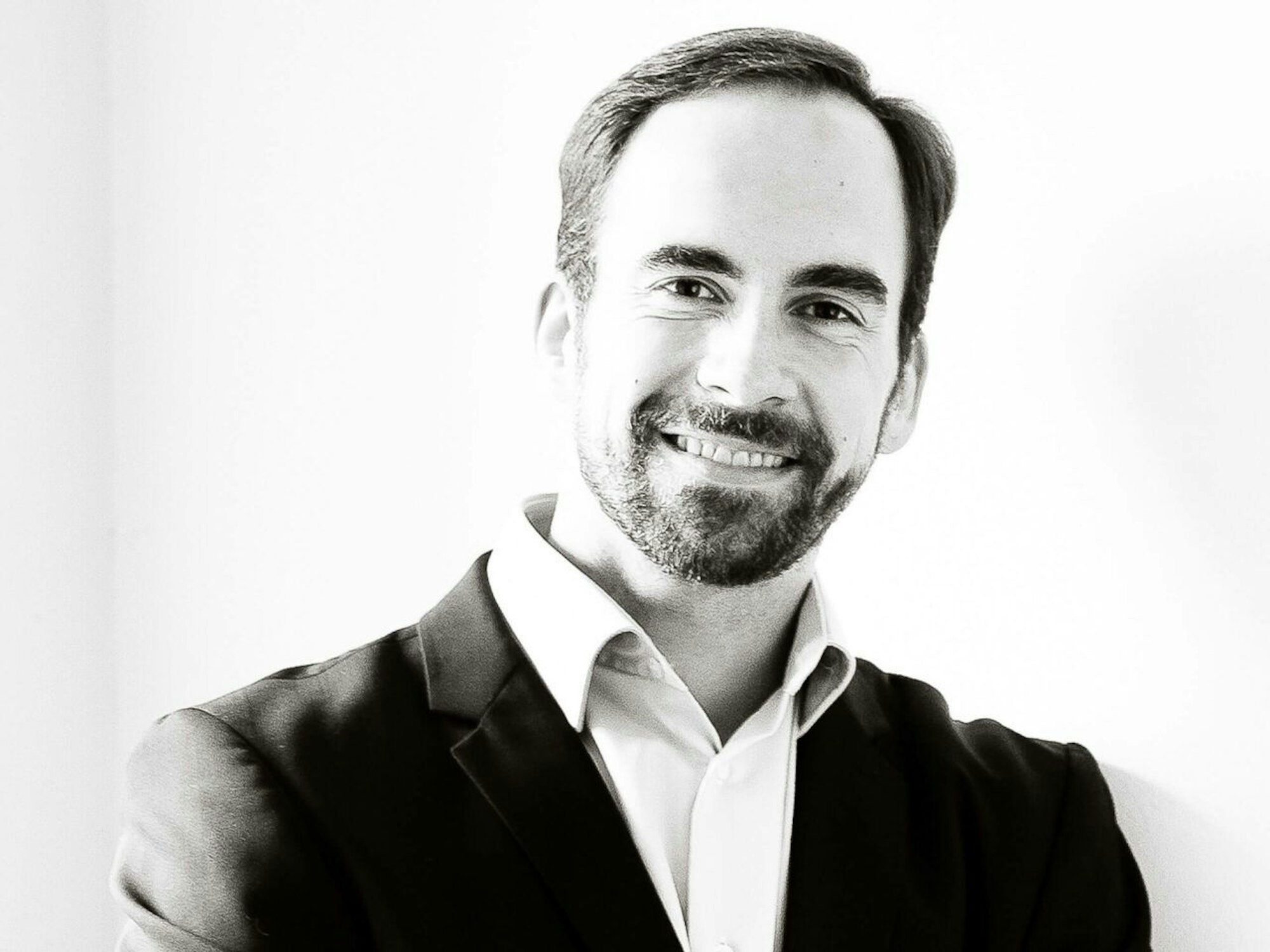 Ein bärtiger Mann mit Anzug und Hemd lacht in die Kamera. Das Foto wurde in schwarz-weiß aufgenommen