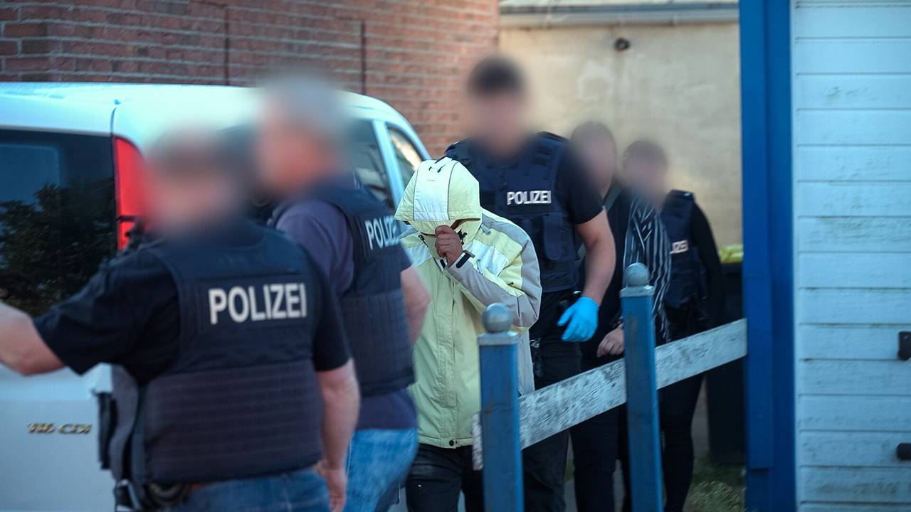 Polizeibeamte, deren Gesichter unkenntlich gemacht worden sind, führen einen Tatverdächtigen ab. Der Mann hat die Kapuze seines gelben Anoraks über das Gesicht gezogen.
