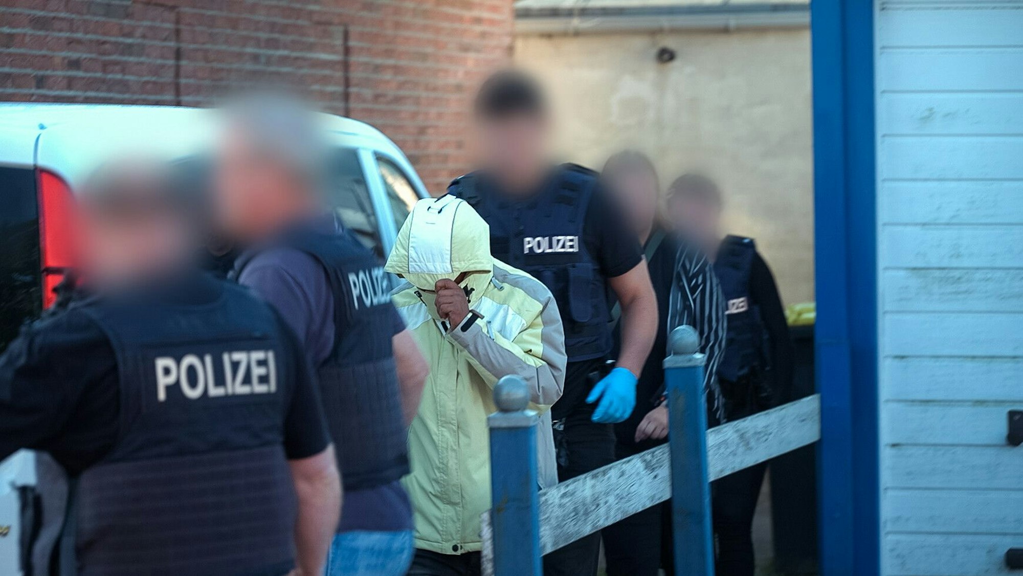 Polizeibeamte, deren Gesichter unkenntlich gemacht worden sind, führen einen Tatverdächtigen ab. Der Mann hat die Kapuze seines gelben Anoraks über das Gesicht gezogen.