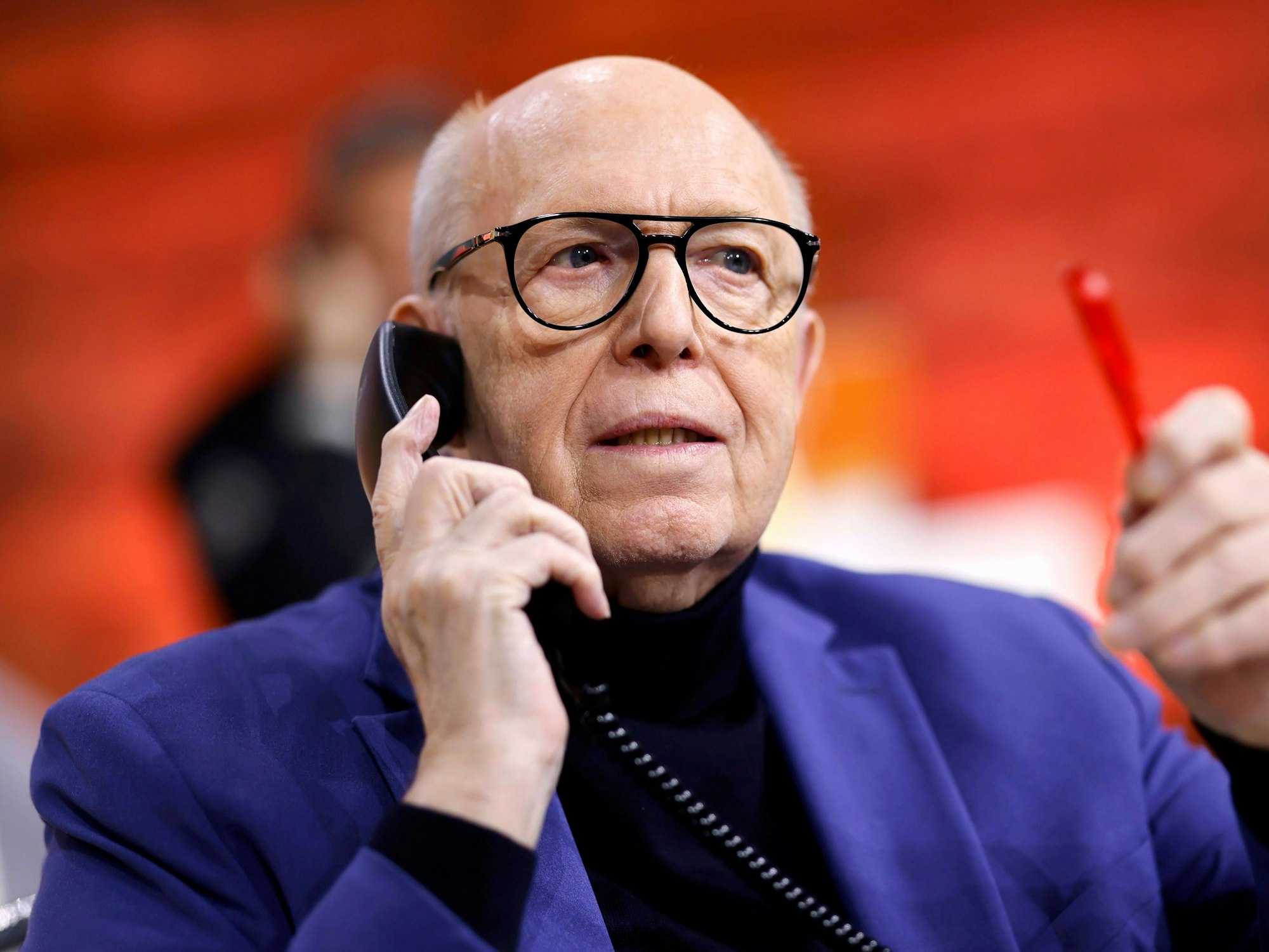 Reiner Calmund hält mit der rechten Hand ein schwarzes Telefon an sein Ohr. In der linken Hand hält er einen roten Kugelschreiber.