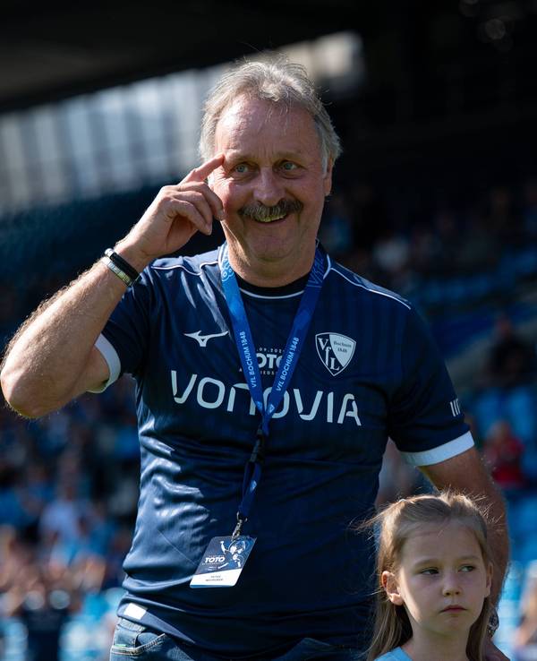Peter Neururer trägt ein Trikot des VfL Bochum und fasst sich mit dem rechten Zeigefinger an seine Augenbraue.