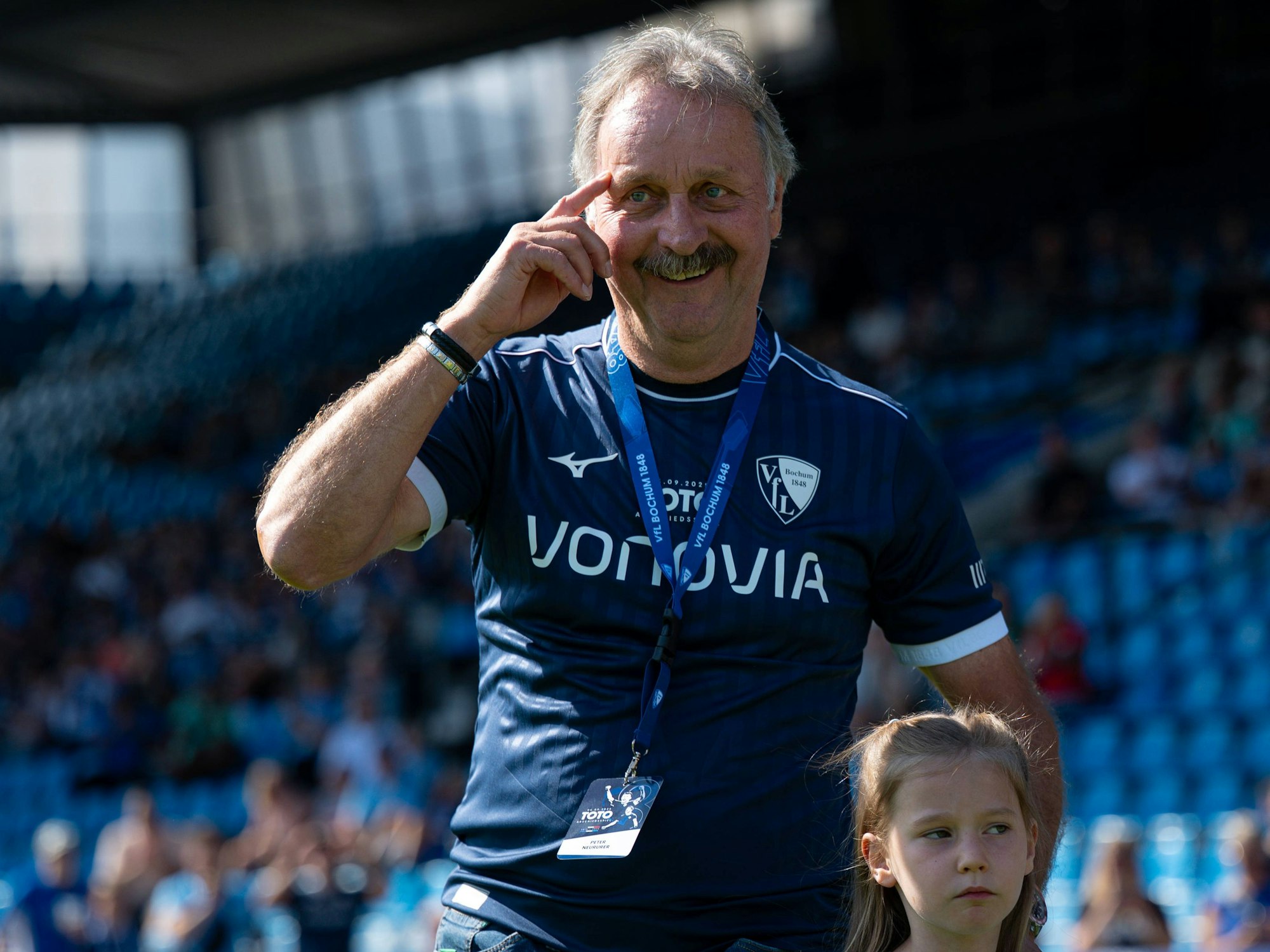 Peter Neururer trägt ein Trikot des VfL Bochum und fasst sich mit dem rechten Zeigefinger an seine Augenbraue.