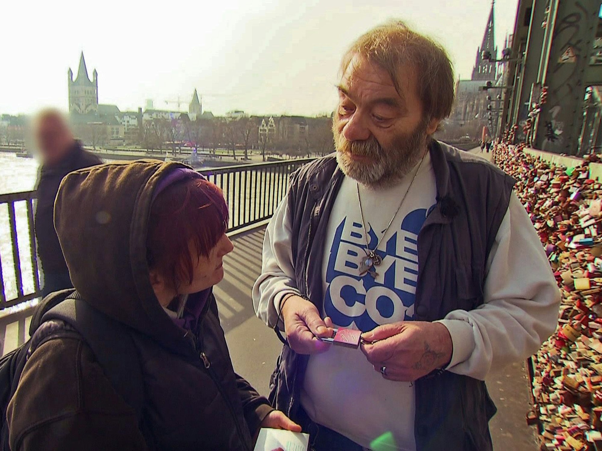 Ein Mann und eine Frau stehen vor den Liebesschlössern auf der Hohenzollernbrücke, er hält ein Liebesschloss in der Hand.