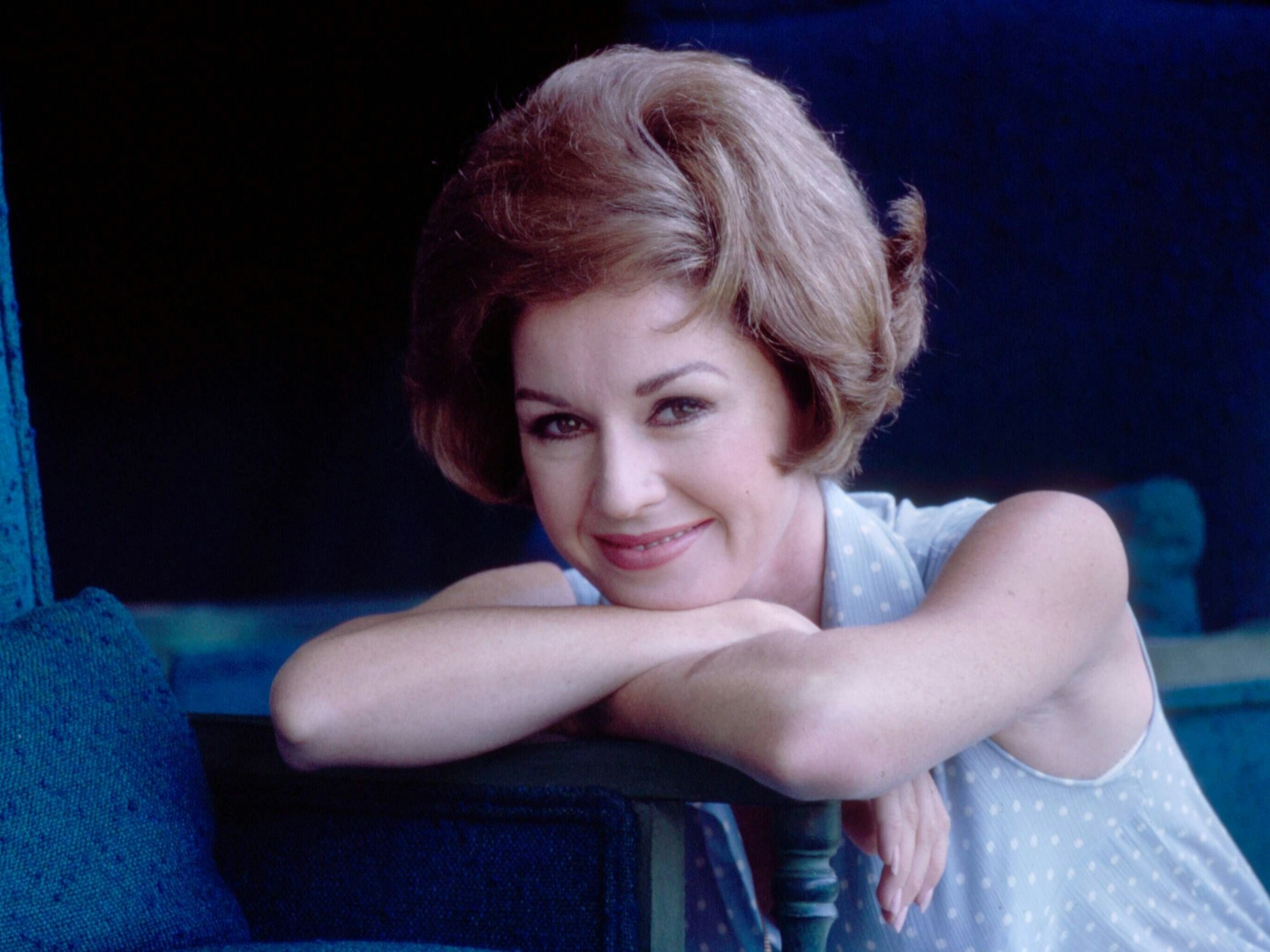 Eine Frau mit auftoupierten Haaren lehnt sich an einen Stuhl.