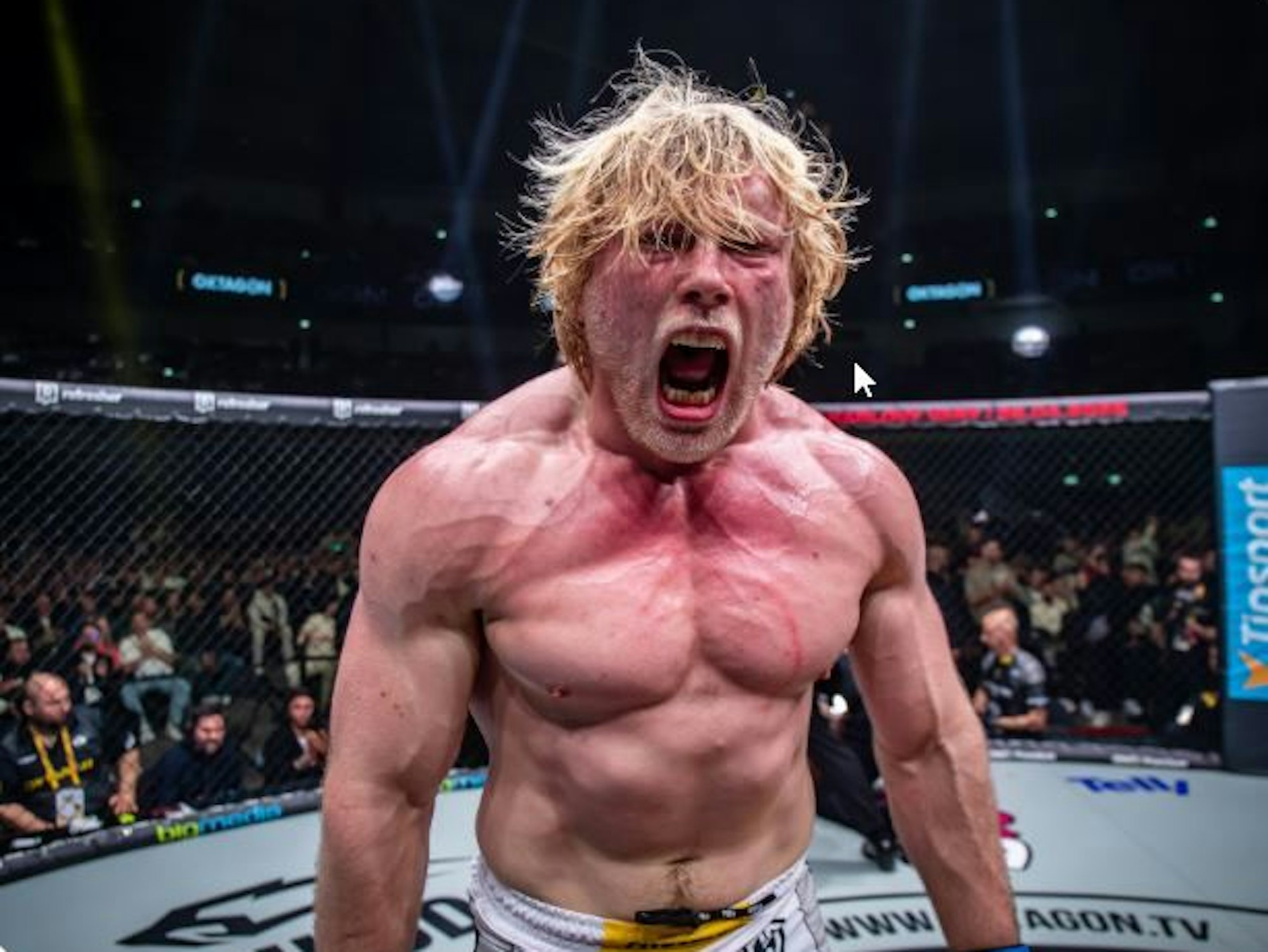 Ein muskelbepackter Mann mit nacktem Oberkörper reißt den Mund zu einem Schrei auf. Er steht in einem Kampfring.