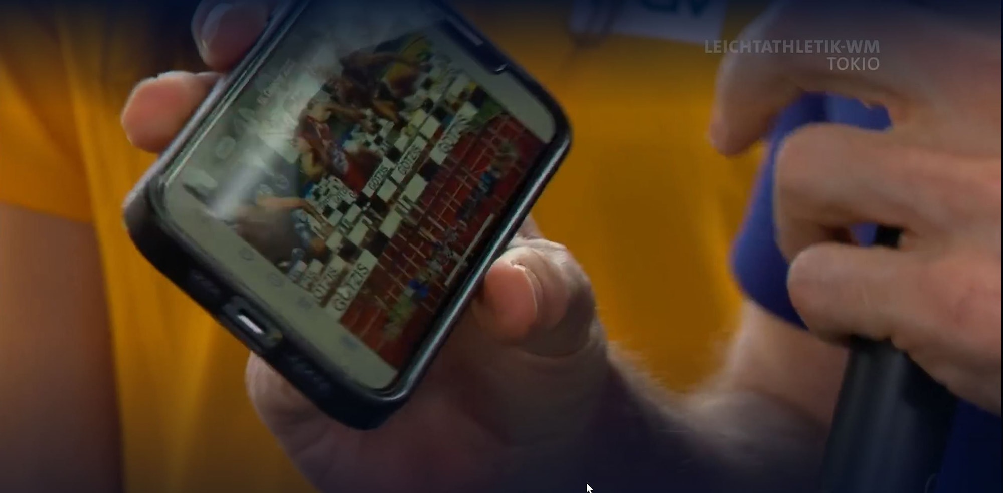 Eine Hand hält ein Handy in die Kamera, auf dem ein Hürdenrennen zu sehen ist.