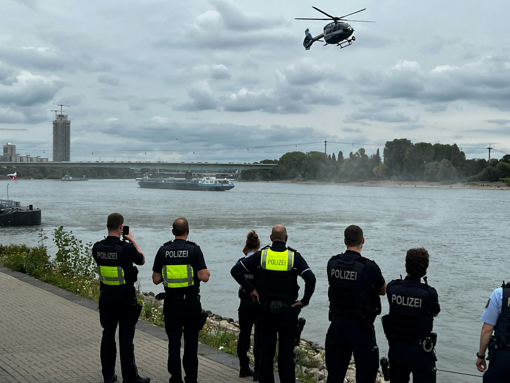 Einsatzkräfte stehen am Rheinufer, über dem Rhein fliegt ein Hubschrauber.