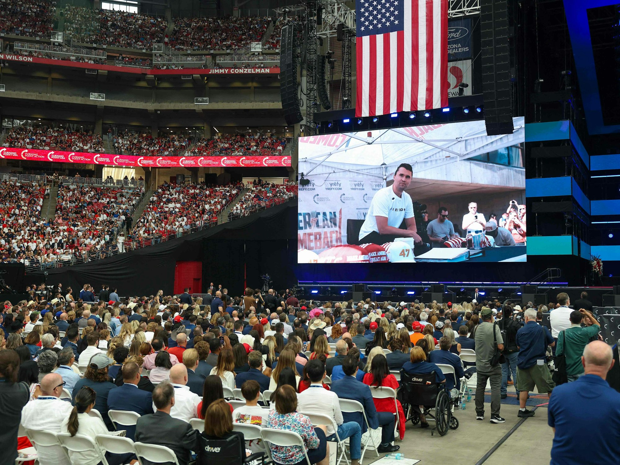 Ein Stadion ist vollbesetzt mit Menschen, alle gucken auf eine große Leinwand.