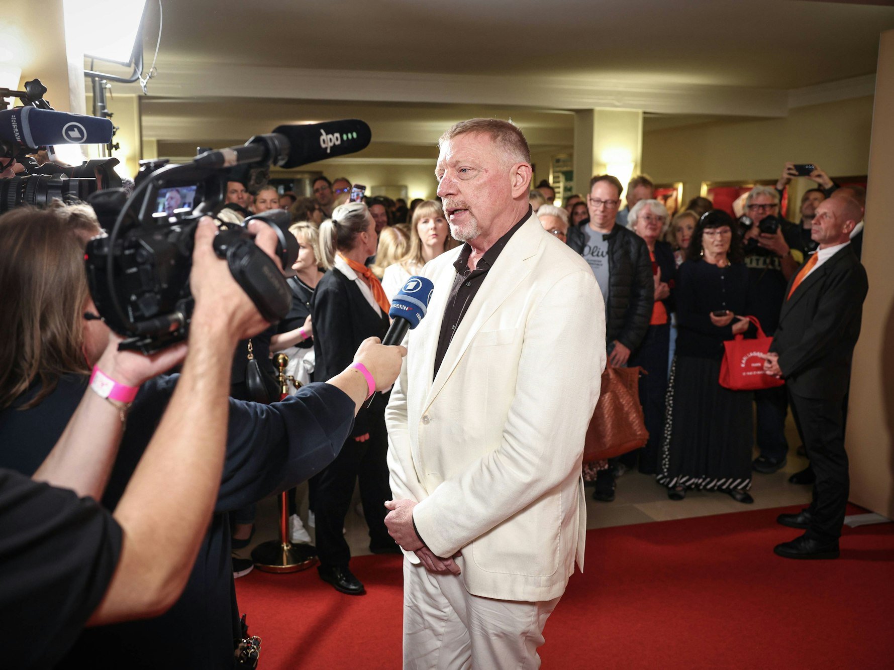 Boris Becker trägt einen weißen Anzug und gibt auf dem roten Teppich ein Interview und wird dabei von mehreren Kameras gefilmt.