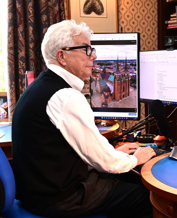 Ein weißhaariger Mann im weißen Hemd mit schwarzer Weste sitzt an einem Schreibtisch voller Bücher und tippt etwas in einen Computer.