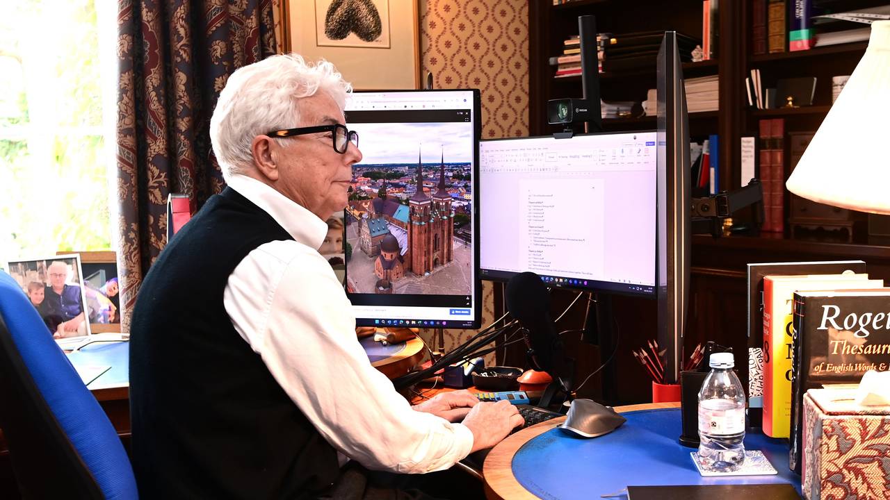 Ein weißhaariger Mann im weißen Hemd mit schwarzer Weste sitzt an einem Schreibtisch voller Bücher und tippt etwas in einen Computer.