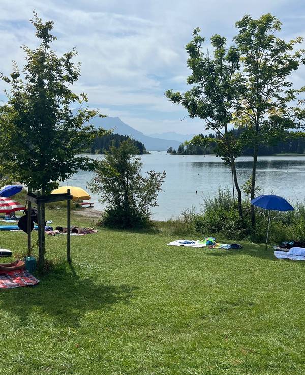 Sonnenschirme auf einer Liegewiese an einem See vor einem atemberaubenden Bergpanorama.