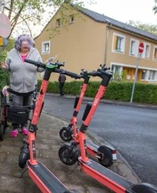 Eine Frau mit Rollator steht vor drei E-Scootern, die den Gehweg blockieren.