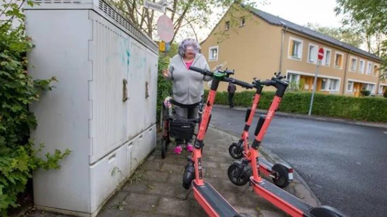 Eine Frau mit Rollator steht vor drei E-Scootern, die den Gehweg blockieren.