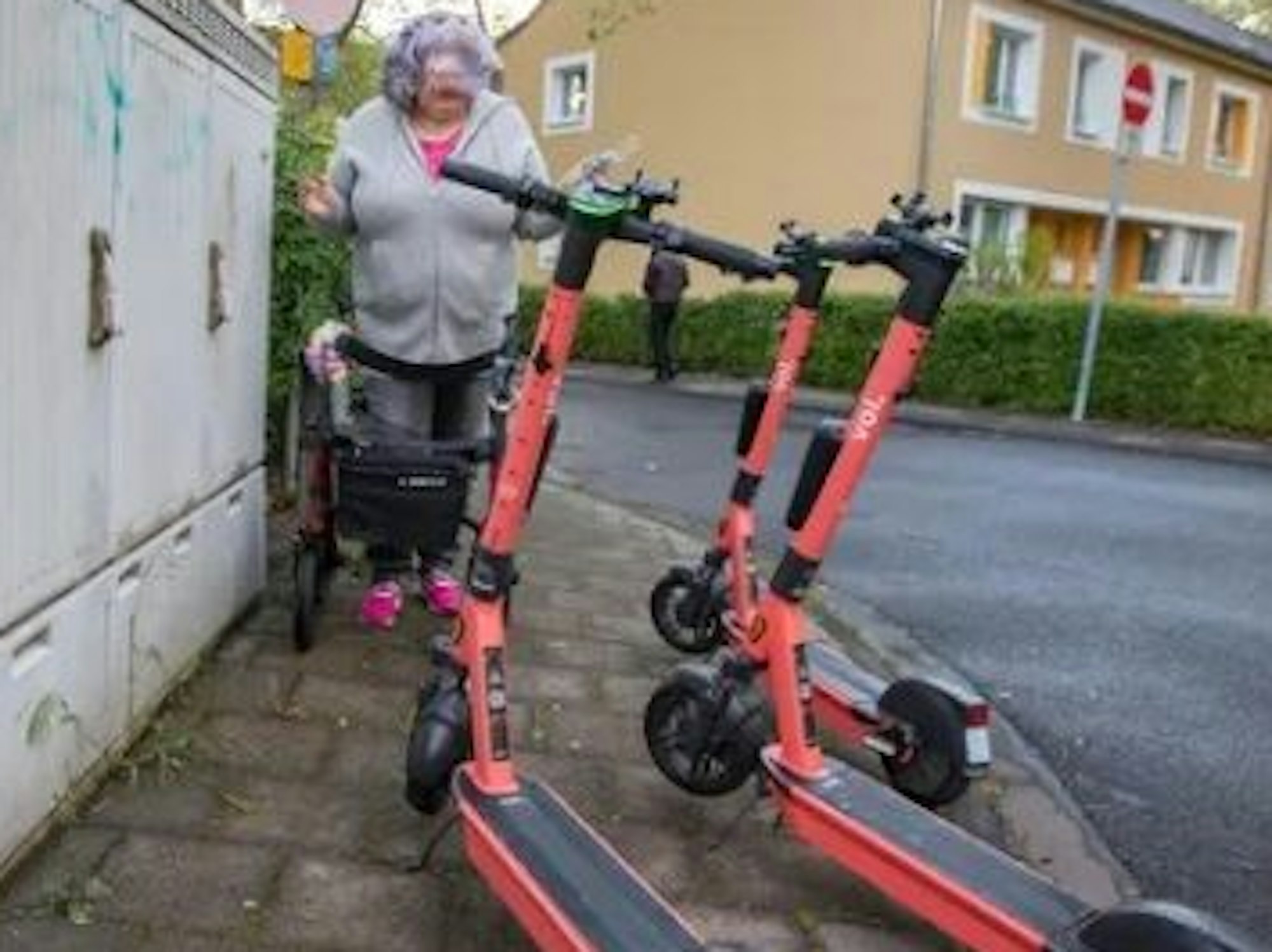 Eine Frau mit Rollator steht vor drei E-Scootern, die den Gehweg blockieren.