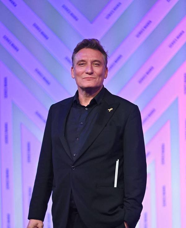 Schauspieler Oliver Masucci steht mit freundlicher Miene vor einer lila-magenta-getönten Wand. Er trägt schwarzes Sakko und schwarzes Hemd.