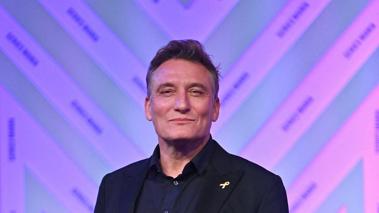 Schauspieler Oliver Masucci steht mit freundlicher Miene vor einer lila-magenta-getönten Wand. Er trägt schwarzes Sakko und schwarzes Hemd.