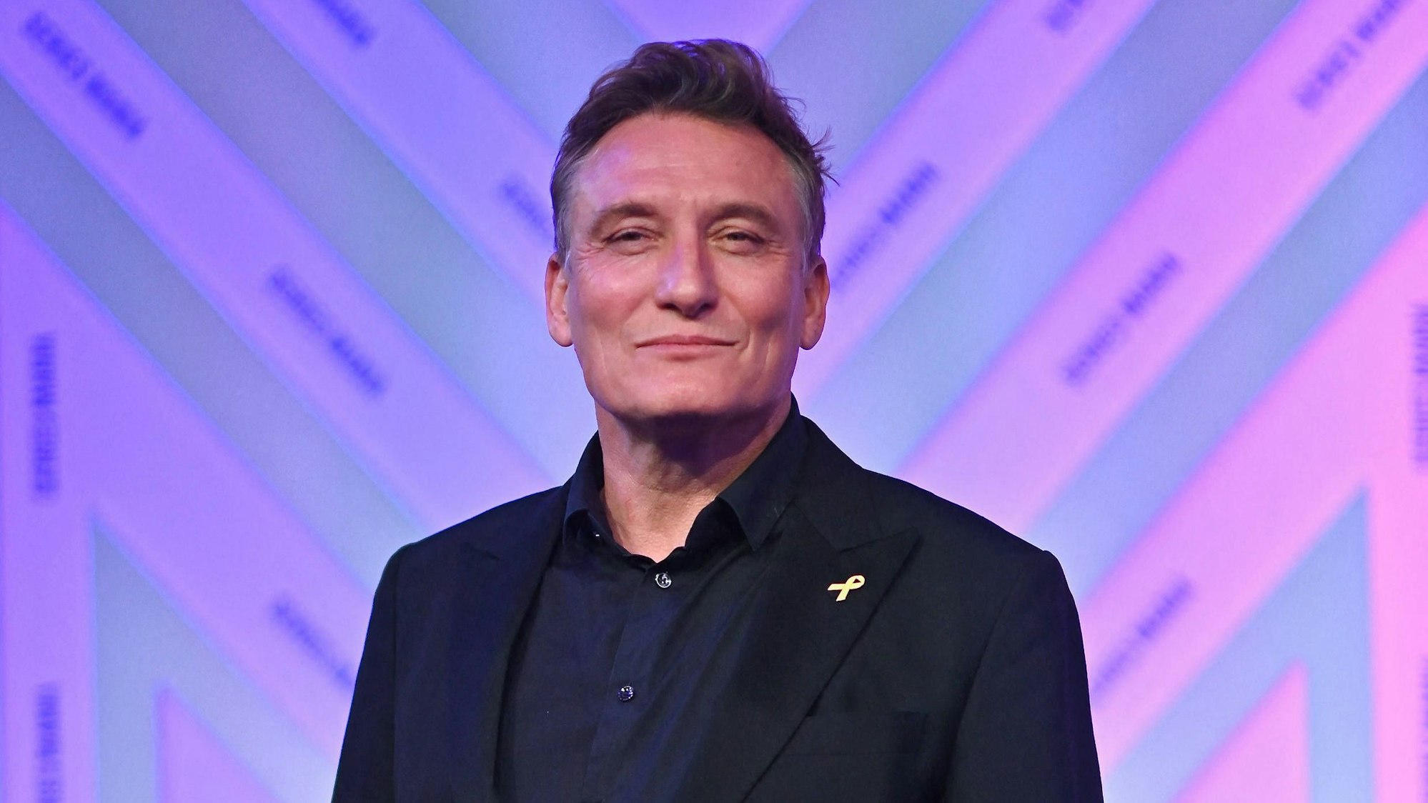 Schauspieler Oliver Masucci steht mit freundlicher Miene vor einer lila-magenta-getönten Wand. Er trägt schwarzes Sakko und schwarzes Hemd.