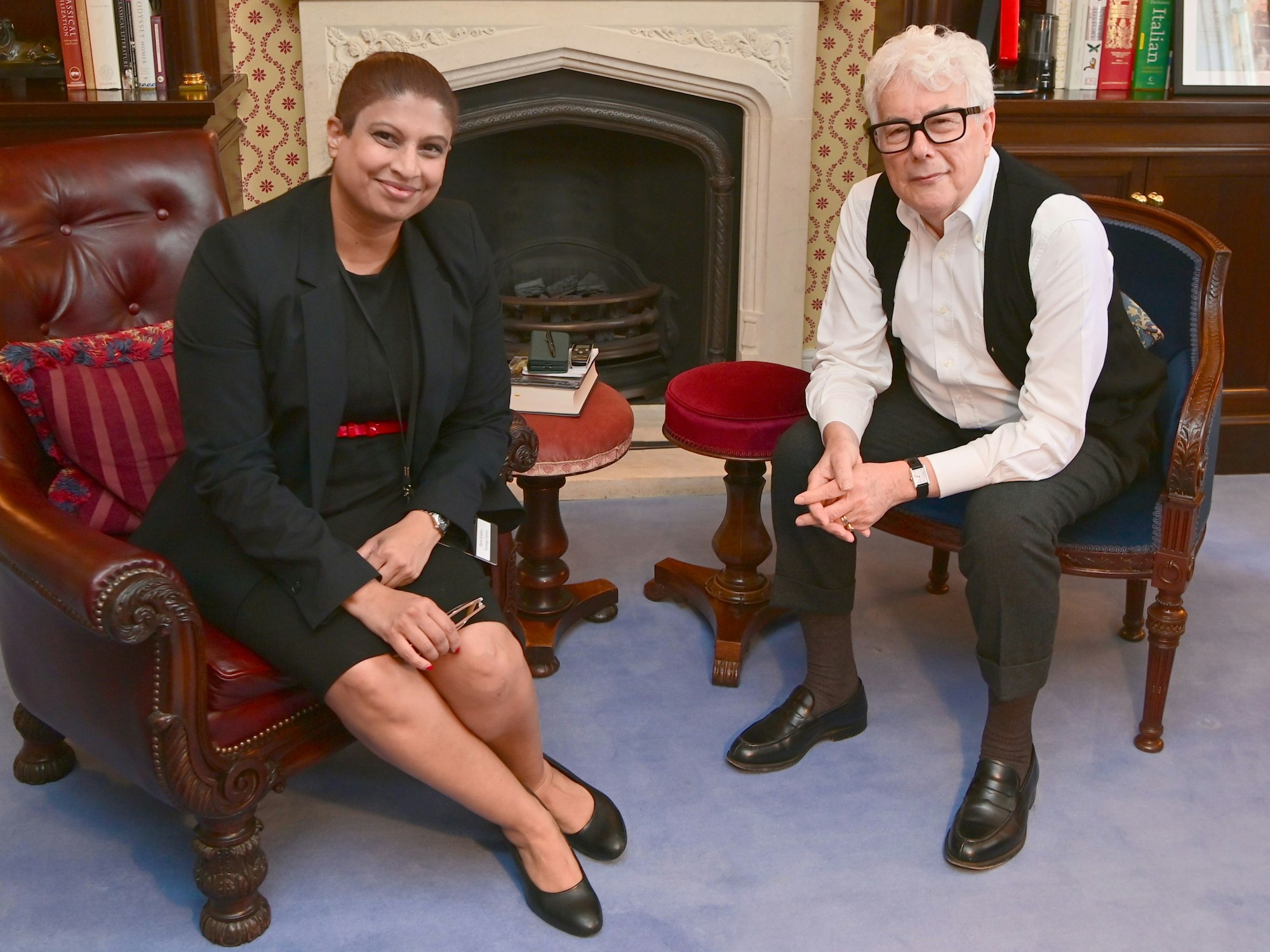 Eine Frau in schwarzem Business-Kostüm und ein Mann mit weißem Haar und offener Anzugweste sitzen vor einem Kamin und unterhalten sich.