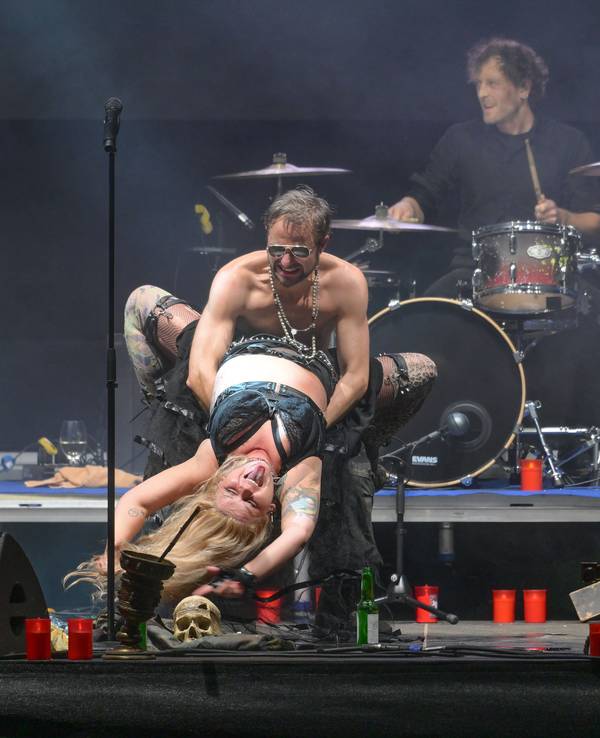 Ein halbnackter Mann mit verspiegelter Brille hält eine tätowierte Frau mit um seine Taille geschlungenen Beinen fest. Im Hintergrund ein Schlagzeug.