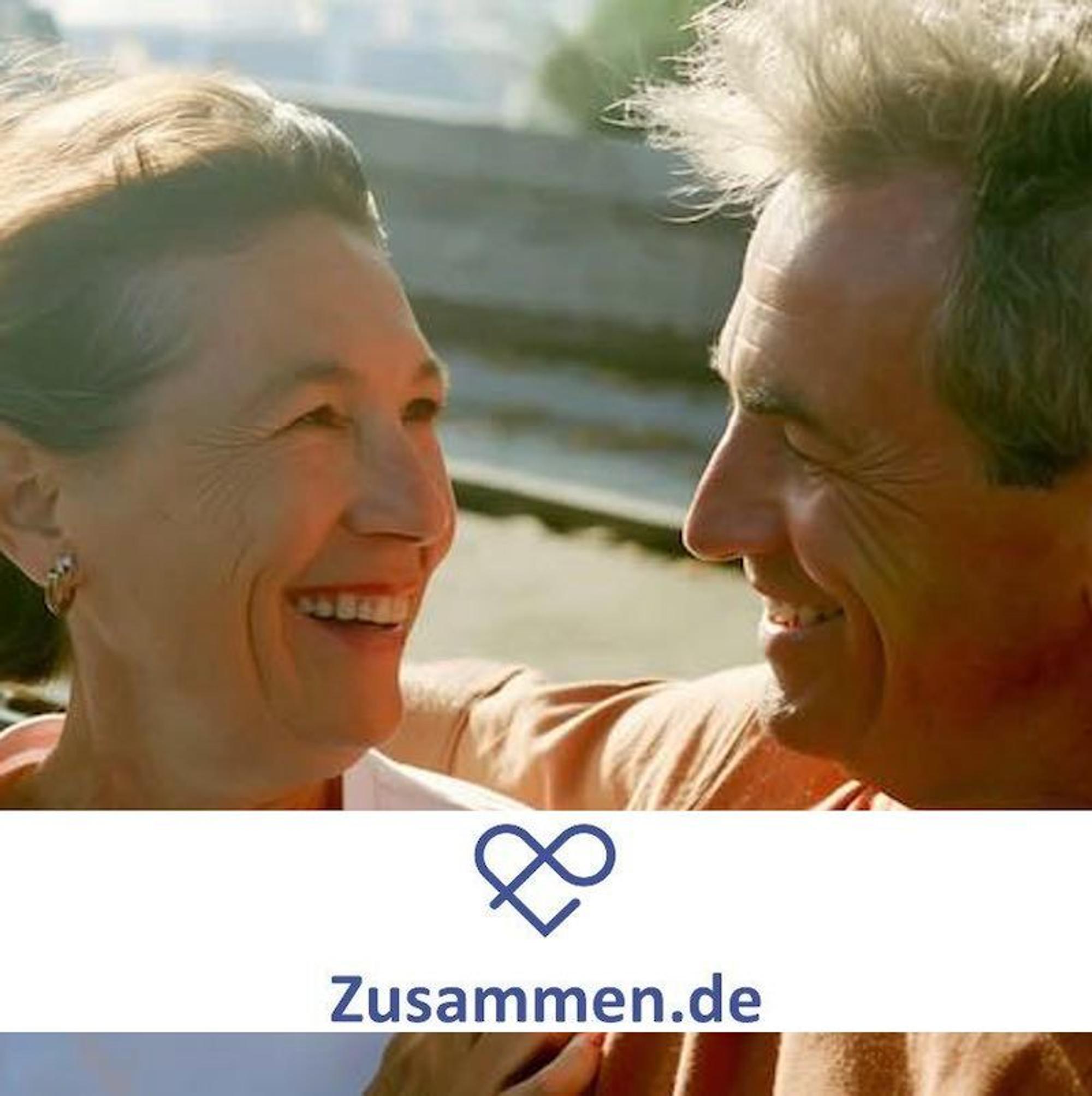 Eine ältere Dame und ein älterer Herr stehen im Freien, halten sich im Arm und lächeln sich ins Gesicht.