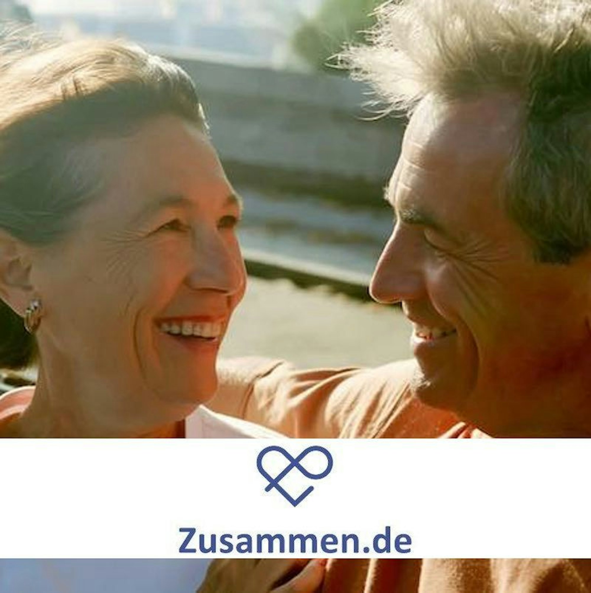 Eine ältere Dame und ein älterer Herr stehen im Freien, halten sich im Arm und lächeln sich ins Gesicht.