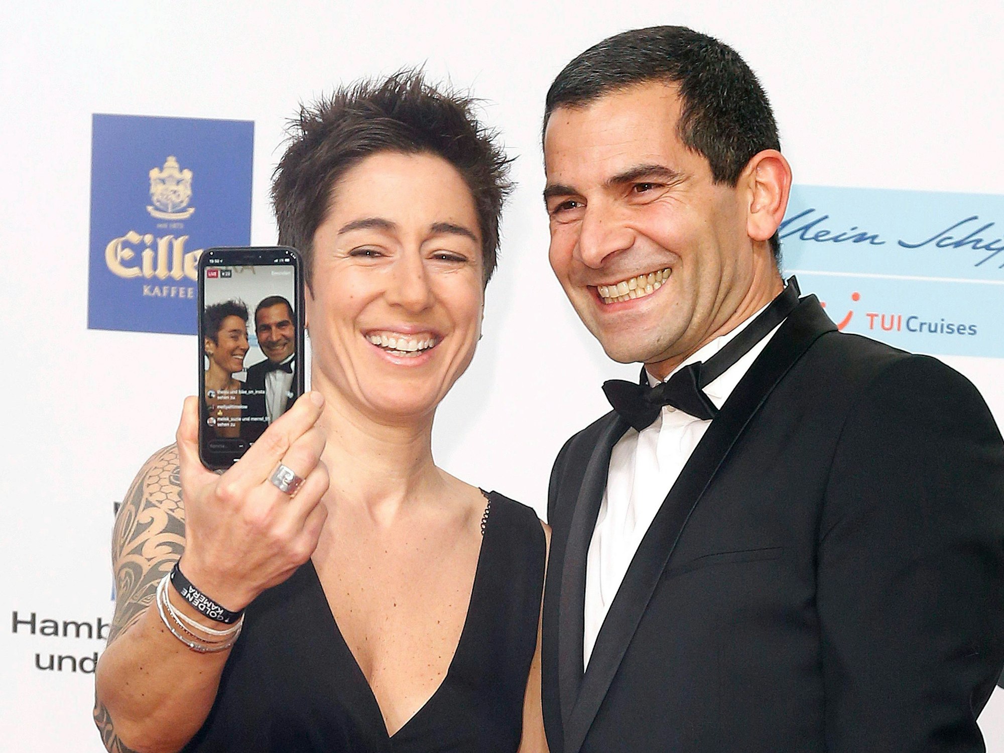 Eine Frau mit kurzen Haaren im Abendkleid hält lachend ein Handy mit einem Selfie von sich und dem Mann hoch, der lachend neben ihr steht.