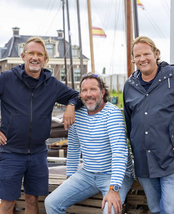 Drei maritim gekleidete Männer stehen am Steuerrad eines Bootes und lachen.