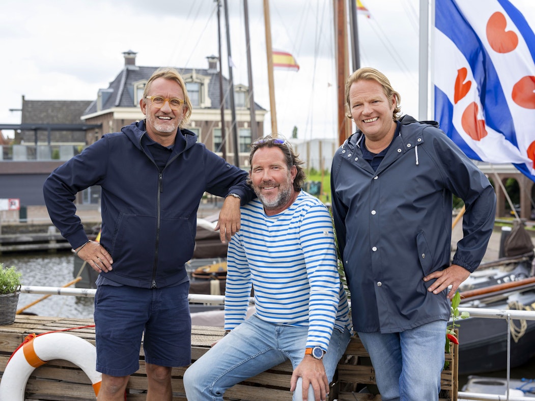 Drei maritim gekleidete Männer stehen am Steuerrad eines Bootes und lachen.