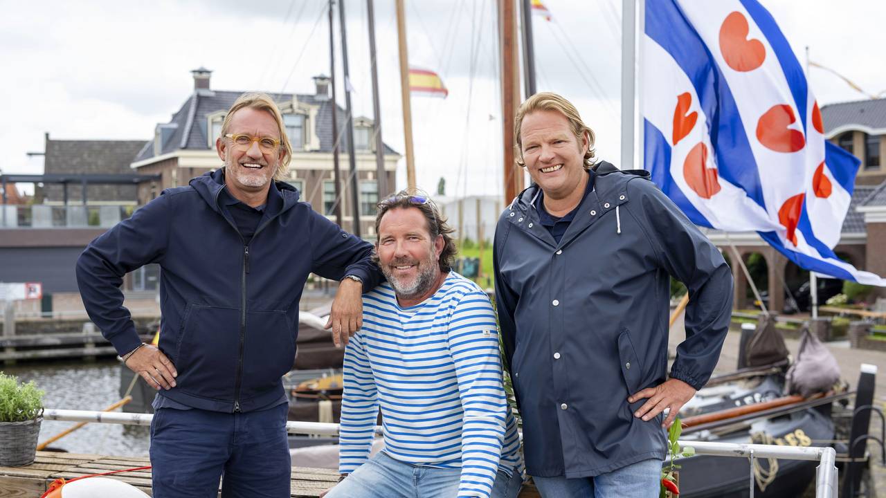 Drei maritim gekleidete Männer stehen am Steuerrad eines Bootes und lachen.