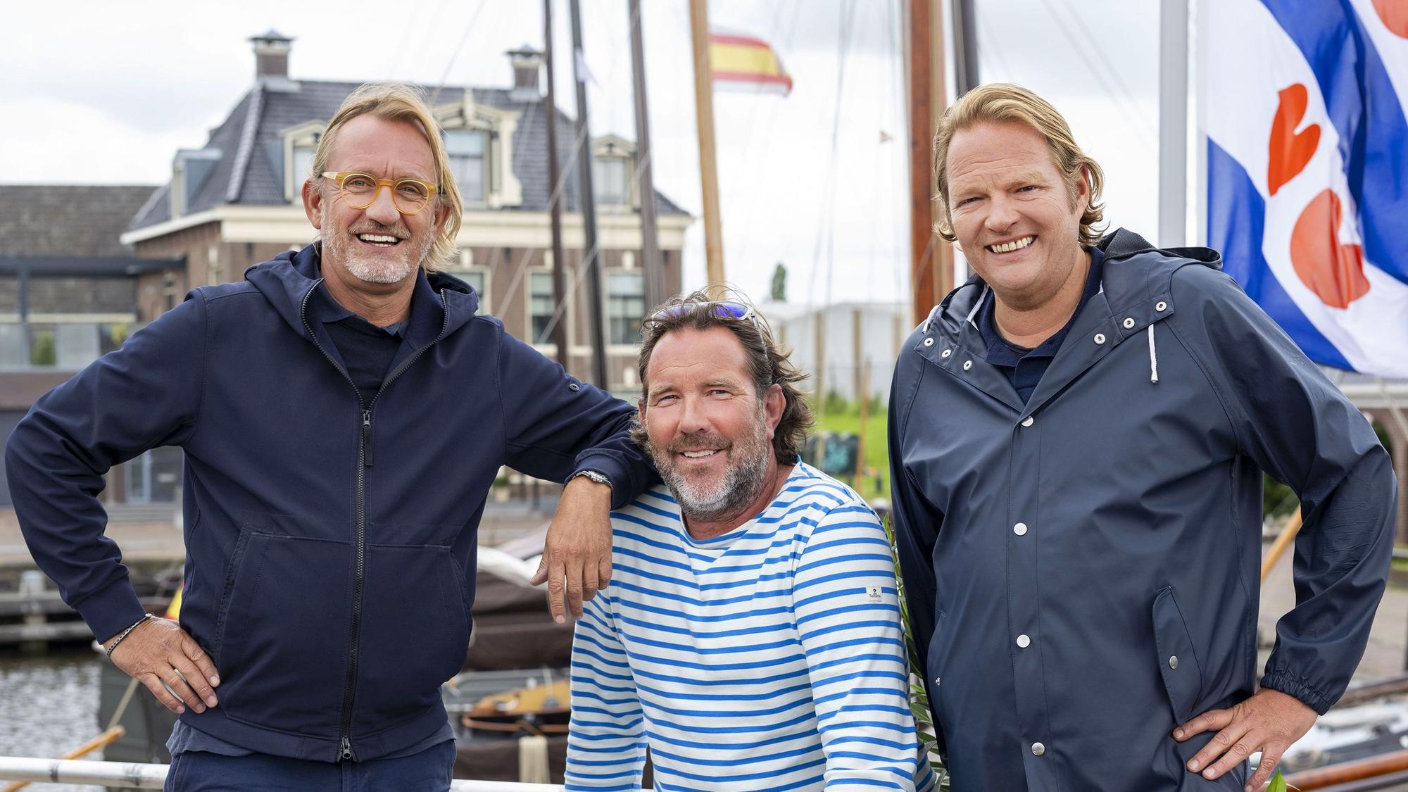 Drei maritim gekleidete Männer stehen am Steuerrad eines Bootes und lachen.