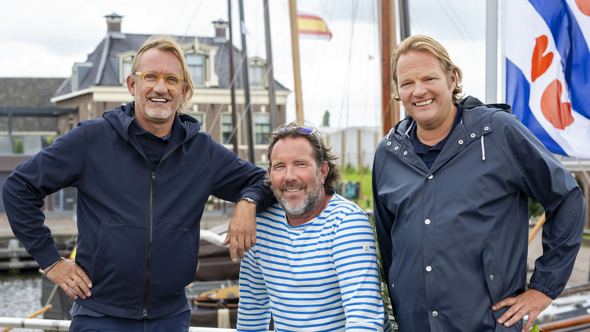 Drei maritim gekleidete Männer stehen am Steuerrad eines Bootes und lachen.