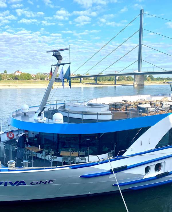 Ein Kreuzfahrtschiff liegt vertäut am Rheinufer, im Hintergrund eine gewaltige Autobahnbrücke.