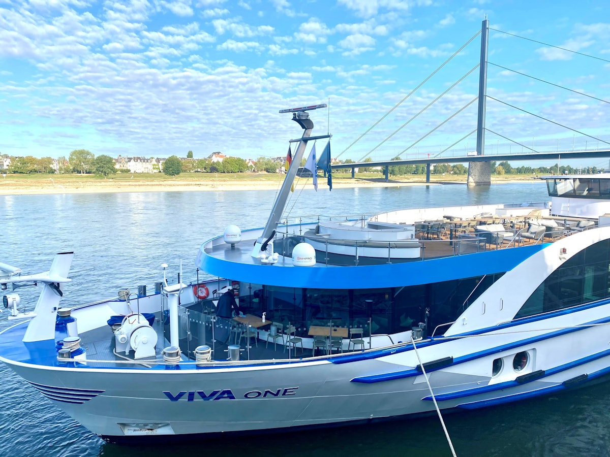 Ein Kreuzfahrtschiff liegt vertäut am Rheinufer, im Hintergrund eine gewaltige Autobahnbrücke.