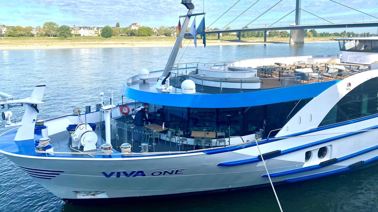 Ein Kreuzfahrtschiff liegt vertäut am Rheinufer, im Hintergrund eine gewaltige Autobahnbrücke.